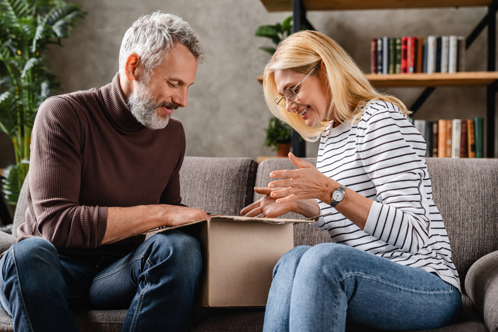 An older couple sitting on a gray couch, smiling and looking at a flat box package placed on their laps in a cozy living room setting.